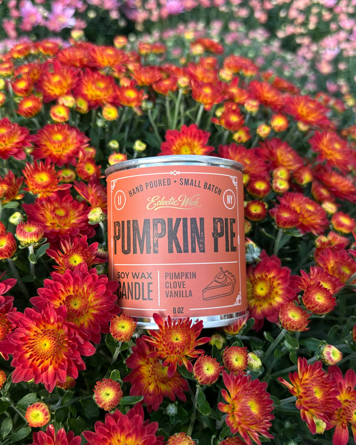 A tin of 'Pumpkin Pie' scented soy wax candle by Eclectic Wick, nestled in a dense bed of red, orange, and yellow chrysanthemums.